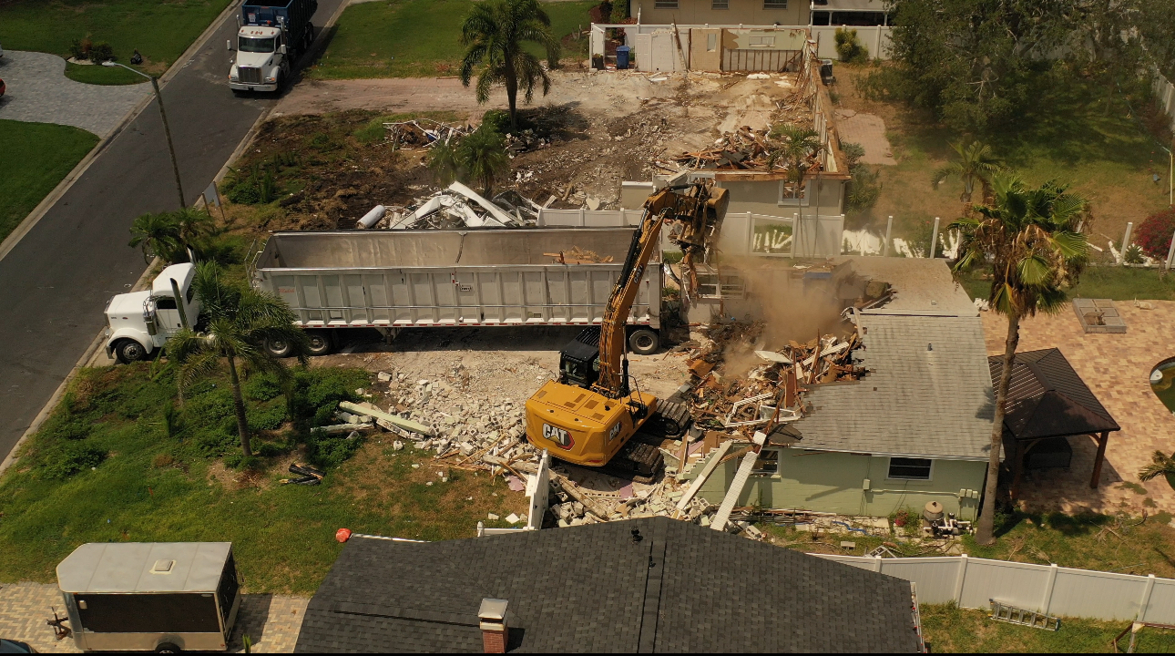 Aerial view of a CAT excavator demolishing a house with a dump trailer on site
